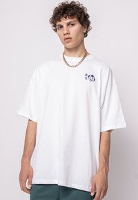 White oversized cotton t-shirt featuring a blue graphic design and text on the left chest. Short sleeves and a crew neckline.
