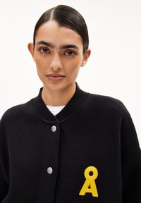 Zwarte cardigan met een geribde kraag, voorzien van zilveren drukknoopjes en een gele geborduurde logo aan de linkerkant. Glad textuur.