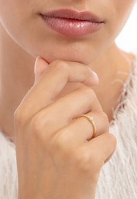 Gold ring with a small clear stone, smooth texture, simple band, worn on a hand with fingers resting under the chin, neutral background.