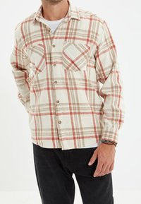 Camisa de franela a cuadros en crema, rojo y beige. Cuenta con dos bolsillos en el pecho, cierre de botones al frente y cuello puntiagudo. Textura suave.