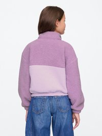 Lavendel fleecetrøje med høj krave, der har et lysere lavendelpanel ved taljen og brede ærmer, parret med blå jeans.