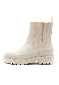 Stivale beige alla caviglia con pannelli laterali elasticizzati, suola testurizzata e una piattaforma bianca maculata. Presenta un testo con il logo sul lato e un manico per il sollevamento.