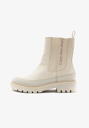 Stivale beige alla caviglia con pannelli laterali elasticizzati, suola testurizzata e una piattaforma bianca maculata. Presenta un testo con il logo sul lato e un manico per il sollevamento.