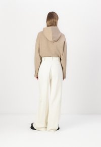 Beige hoodie met een achternaad en capuchon, gecombineerd met witte broek met wijde pijpen, zijzakken en een hoge taille. Zwarte schoenen.