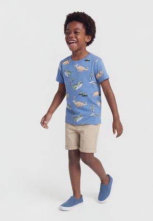 Niño sonriente caminando con una camiseta azul con estampado de dinosaurios, pantalones cortos beige y zapatos azules tipo slip-on sobre un fondo blanco liso.