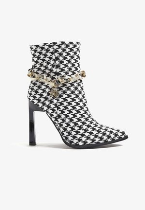 Czarno-białe botki w kratę houndstooth sięgające kostki z wyciągniętym toe, wysokim obcasem szpilką i złotym akcentem w postaci łańcucha z logotypem marki.