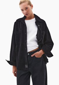 Czarna kordurowa oversize'owa kurtka koszulowa z kołnierzem, z przodem zapinanym na guziki, dwoma kieszeniami na piersi i ribbową teksturą, połączona z czarnymi dżinsami.