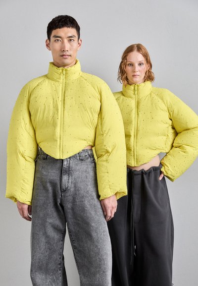 Des vestes doudounes jaunes coupées avec de grandes manches, présentant une texture lisse et des éclats de paillettes subtils, associées à des bas gris et noirs.