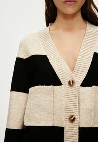 Beige and black striped cardigan featuring a deep V-neck, ribbed collar, two front pockets, and round brown buttons. Textured knit material.
