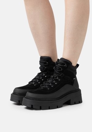 Black ankle boots featuring a combination of suede and synthetic materials, with a thick platform sole and metal eyelets for laces.
