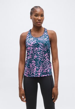 Atletyczna kobieta stojąca, ubrana w różowo-niebieski sportowy top bez rękawów z wzorem oraz czarne legginsy na jednolitym tle.