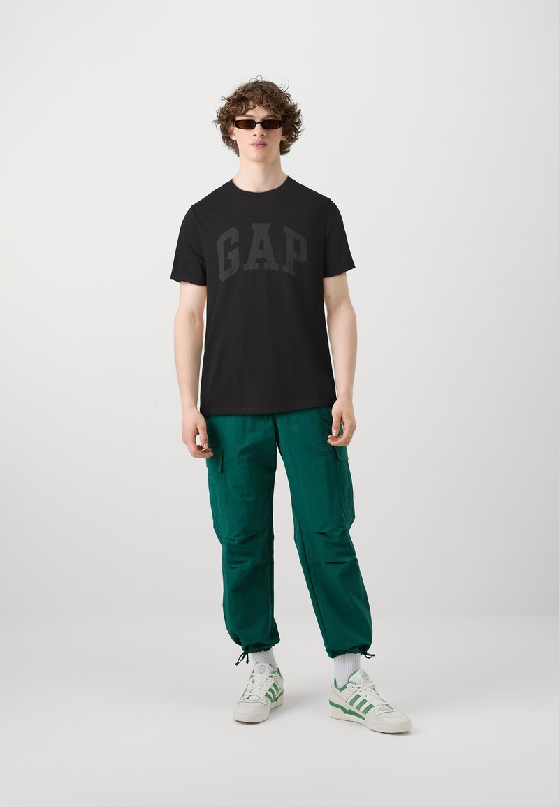 T-shirt noir avec le logo "GAP", associé à un pantalon cargo vert. La tenue est complétée par des baskets blanches avec des accents verts.