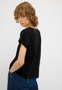 Top corto nero con maniche a giro e chiusura a zip visibile sul retro. Indossato con jeans blu caratterizzati da decorazioni con zip.