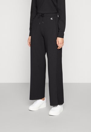 Person wearing black wide-leg pants with drawstring waist and small CK logo, paired with white platform sneakers on gray floor.