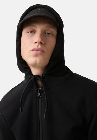 Schwarze Zip-Hoodie mit Kapuze und Kordelzug, ausgestattet mit einem kleinen Emblem und einem strukturierten Stoff. Eine passende schwarze Kappe wird darüber getragen.