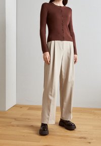 Bruine geribbelde cardigan met lange mouwen en knopen, gecombineerd met beige broek met wijde pijpen en donkere schoenen met een stevige hak. Achtergrond van houten vloer.