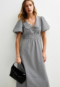 Robe longue à carreaux noir et blanc avec des manches bouffantes, un col froncé et un détail à nouer au niveau de la poitrine ; assortie à un sac noir.