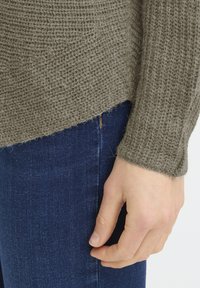 Pull en tricot kaki avec un motif texturé et des poignets côtelés, associé à un jean en denim bleu. Le pull présente un ourlet droit.