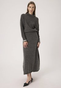 Femeie îmbrăcată cu o rochie tricotată lungă de culoare gri cărbune, cu mâneci lungi și tocuri negre ascuțite, stând în fața unui fundal deschis simplu.