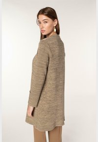 Beige texturerad kappa med skräddarsydd passform, långa ärmar och sidofickor. Bärs över beige byxor med en enkel design och ren siluett.