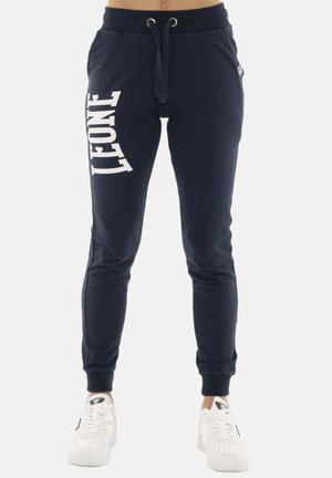 Pantalon de jogging bleu marine avec "LEONE" imprimé verticalement sur la cuisse gauche, porté avec des baskets blanches. Taille à cordon et chevilles côtelées.