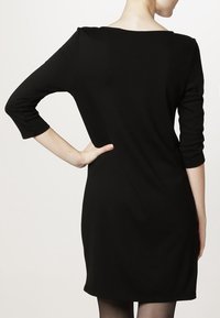 Femme portant une robe noire jusqu'aux genoux avec des manches trois-quarts, vue de derrière avec une main sur la hanche.