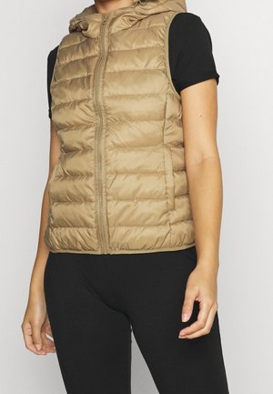 Bezrekawnik w jasnobrązowym kolorze z pikowaniem, z przednim zamkiem błyskawicznym i kapturem, noszony na czarnej koszulce z krótkim rękawem i czarnych spodniach.