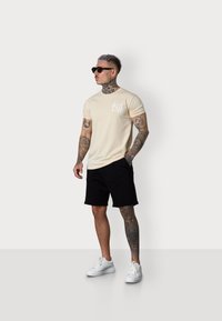 Sleeved beige t-shirt with a white graphic, paired with black shorts and white sneakers. Model wears black sunglasses and has visible tattoos.