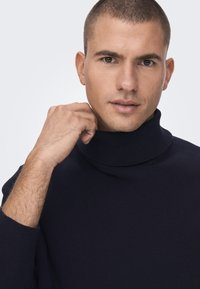 Mornarica rebrasti jumper s visokim ovratnikom. Tekstura tkanine čini se mekanom, s prilagodljivim dizajnom i suptilnim rastezanjem. Ruka postavljena na ovratniku.