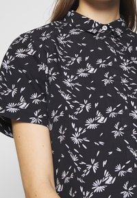 Camisa negra de manga corta con patrón floral blanco, cuello abotonado y tejido suave. Presenta un corte relajado y una textura sutil.