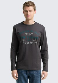 Dunkelgraues Langarm-T-Shirt mit Rundhalsausschnitt, mit türkisfarbener Druckschrift. Getragen mit blauen Jeans, minimales Design, glatte Textur.