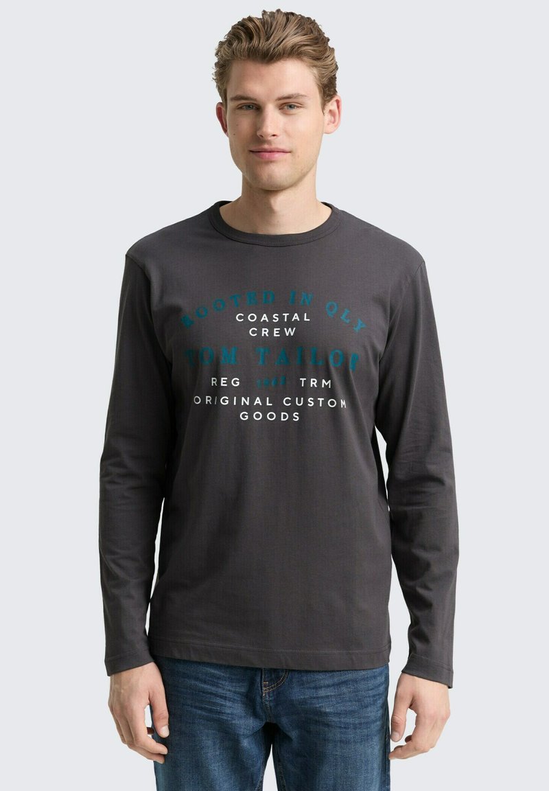 Dunkelgraues Langarm-T-Shirt mit Rundhalsausschnitt, mit türkisfarbener Druckschrift. Getragen mit blauen Jeans, minimales Design, glatte Textur.