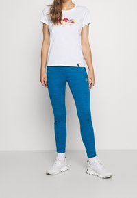 White t-shirt with colorful mountain graphic, paired with blue ribbed leggings. Model wears white sneakers, standing on a plain background.