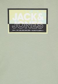 Camiseta verde claro con un logotipo rectangular negro y amarillo que lleva impreso en grandes letras blancas "JACK & JONES" y las coordenadas debajo.