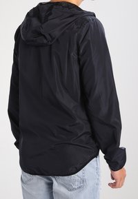 Svart lättviktsjacka med huva, slät yta, elastiska ärmslut och en lätt rundad fåll. Tillverkad av vattentåligt material.