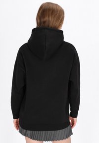 Fekete kapucnis pulóver puha anyagból, kapucnival és laza szabással, szürke pliszírozott szoknya fölött viselve. Látható varrások és mandzsetták.