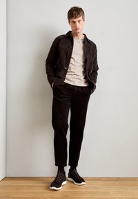 Dark corduroy jacket over a light beige T-shirt, paired with tapered black corduroy pants and black sneakers with white soles.