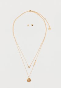 Collana a più strati in tonalità oro, con pendente circolare testurizzato, accento a stella e lettere in blocco; abbinata a orecchini a forma geometrica.