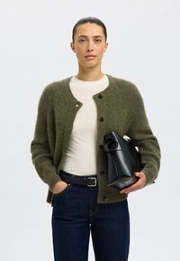 Olívazöld kötött kardigán, kerek nyakkivágással és fekete gombokkal, krémszínű inggel és sötét farmernadrággal párosítva. Fekete táskát tart a kezében.