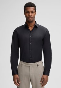 Uomo che indossa una camicia nera attillata con bottoni e pantaloni beige su misura, in piedi contro uno sfondo grigio chiaro uniforme.