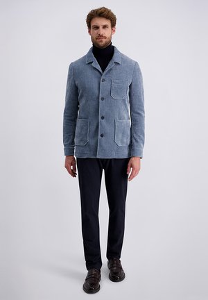 Mann, der eine hellblaue Kordjacke mit Knöpfen und drei Vorderaschen, einen schwarzen Rollkragenpullover, schwarze Hosen und dunkelbraune Schuhe trägt, vor einem weißen Hintergrund steht.