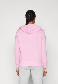 Rosa Kapuzenpullover mit Kordelzug, gerippten Bündchen und Saum. Der Stoff wirkt weich und leicht strukturiert. Kombiniert mit grauen Sweatpants.