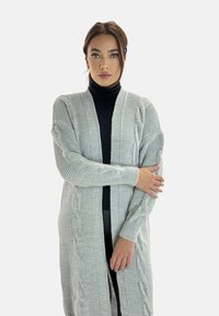 Ljusgrå stickad cardigan med flätmönster, ribbade ärmslut och öppen fram. Har en mjuk textur och avslappnad passform.