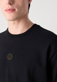 Fekete pamut pulóver, bordázott kerek nyakkivágással, arany színű, stilizált "B" betűvel ellátott szövet folttal a gallér közelében.
