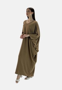 Bruine maxi-jurk met een losse, gedrapeerde silhouette en grote mouwen. Gladde textuur. Gepaard met witte blokhakken sandalen.