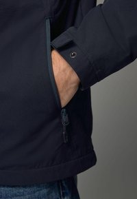 Giacca blu scuro con una texture morbida, dotata di una tasca laterale con zip e polsini regolabili con chiusura a scatto.
