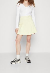 Záhybová žlutá skater sukně vyrobená z lehkého materiálu, kombinovaná s bílým žebrovaným topem s dlouhým rukávem a černobílými teniskami.