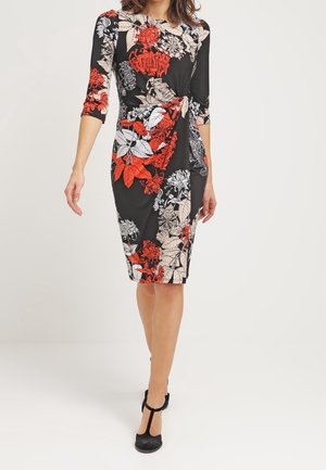 Kobieta ubrana w czarną sukienkę do kolan z czerwonymi, białymi i beżowymi kwiatowymi wzorami, z rękawami do łokcia, oraz czarne sandały typu T-strap, idzie do przodu.