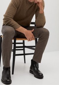 Suéter marrón acanalado, pantalones de cuadros marrones y botas de cuero negras hasta el tobillo, sentada en una silla negra contra un fondo neutro.