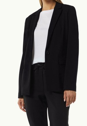 Femme portant un blazer noir, un pantalon noir avec cordon de serrage et un t-shirt blanc à col rond, vue des épaules jusqu'au milieu des cuisses.